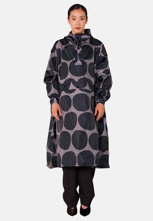 Vrouw staat, draagt een losse zwarte en grijze regenjas met capuchon met grote stippen over een zwarte broek en zwarte schoenen.