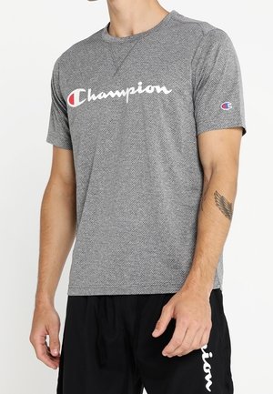 Man draagt een grijs Champion T-shirt met korte mouwen en zwarte korte broek, met een vleugel-tatoeage zichtbaar op zijn linker onderarm.