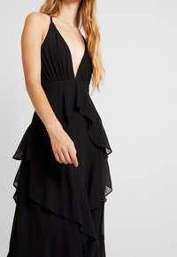 Kobieta ubrana w czarną, bezrękawową sukienkę z głębokim dekoltem w kształcie litery V i warstwowymi falbanami, stojąca na neutralnym tle.