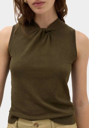 Frau trägt ärmelloses olivgrünes Stricktop mit gedrehtem Detail am Ausschnitt und beige Hose mit Knopfverschluss.