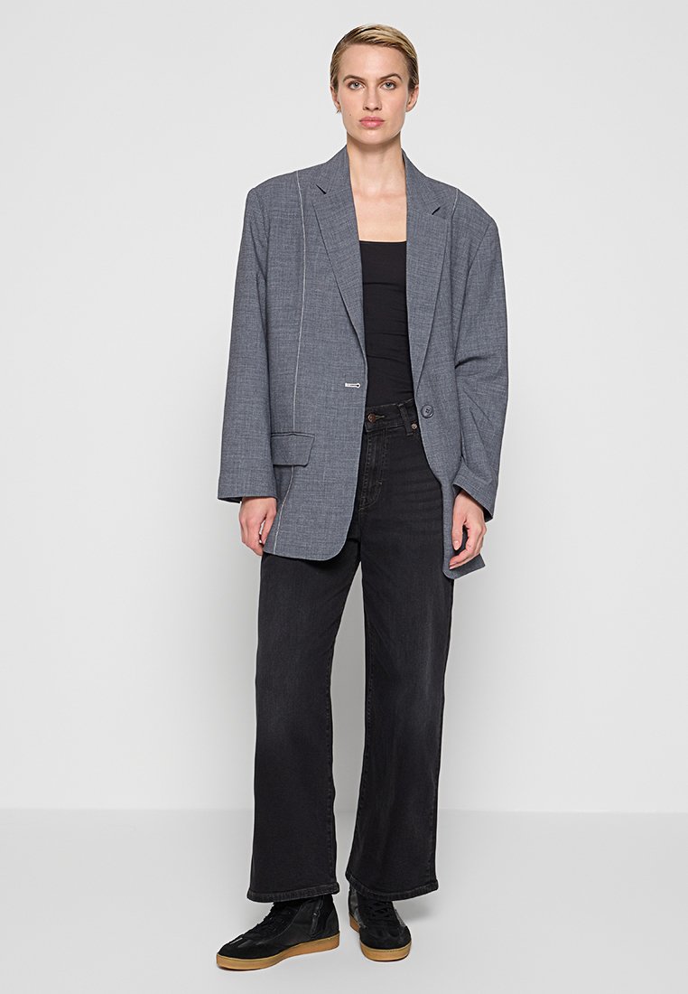 Frau steht vor einem weißen Hintergrund und trägt einen übergroßen grauen Blazer, ein schwarzes Oberteil, weite schwarze Jeans und schwarze Sneaker mit Gummisohlen.