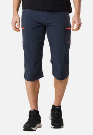 Muž nosí námornícky modré cargo šortky po kolená s oranžovými zipsami a čierne športové topánky s bielymi podrážkami.