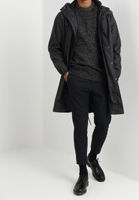 Zwarte waterdichte parka met een capuchon, een grijze gestructureerde trui, slim zwarte broek en zwarte schoenen met veters, tegen een neutrale achtergrond.