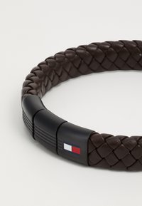Brun lærflettet armbånd med en matt svart lås med en logo i blått, hvitt og rødt. Glatt tekstur med en fleksibel form.