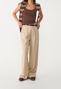 Beige wijde broek met plooien aan de voorkant, gecombineerd met een bruine tanktop en een gestreepte bruine gebreide trui over de schouders gedrapeerd.