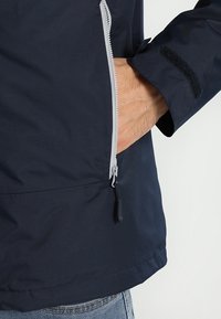 Marinblå vattentät jacka med en ficka med dragkedja, grå dragkedjedetalj och kardborrestängning vid ärmarna. Tyget verkar slätt med en matt yta.