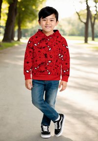 Ragazzino che sorride, indossando un felpa rossa di Spider-Man, jeans blu e sneakers nere, in piedi su un sentiero alberato alla luce del giorno.