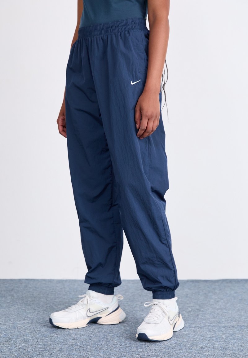 Osoba v tmavě modrých sportovních kalhotách Nike a bílých teniskách Nike stojící na šedém koberci před jednobarevným pozadím.