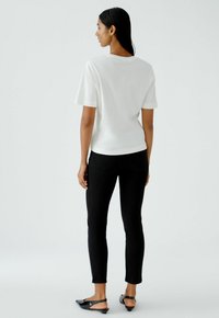 Weißes Kurzarmshirt aus glattem Stoff, kombiniert mit schwarzen, taillierten Hosen und schlichten schwarzen Slingback-Schuhen. Einfarbiger Hintergrund.