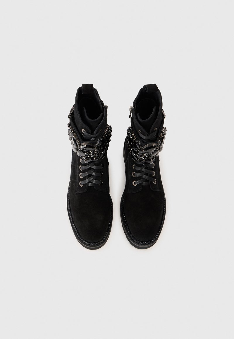 Bottes de cheville en daim noir avec une finition texturée, présentant des accents en métal argenté et des lacets. Le design comprend un détail de chaîne unique autour de la cheville.