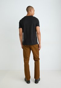 Black short-sleeve t-shirt paired with brown straight-leg trousers and black lace-up boots, seen from the back against a neutral background.