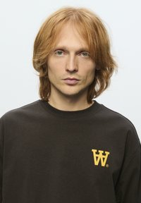 Brauner Pullover aus weichem Stoff mit einem gelben Logo auf der linken Brust. Das Design umfasst einen runden Halsausschnitt und eine lockere Passform.