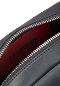 Sort lædertaske med åben sølvlynlås, der afslører rød inderforing med "HUGO"-branding i sorte bogstaver.