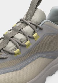 Κοντινό πλάνο ενός γκρι και μπεζ αθλητικού παπουτσιού με γκρι κορδόνια περασμένα μέσα από κίτρινες υφασμάτινες θηλιές, με ετικέτα "DRYVENT" στην πλαϊνή επιφάνεια.