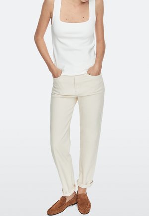Person trägt ein weißes ärmelloses Top, hellbeige hochgekrempelte Hosen und braune Wildleder-Loafer mit Händen in den Taschen.