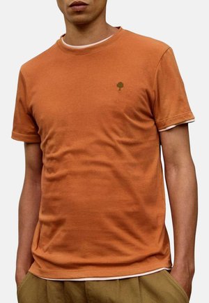 Homme portant un t-shirt à manches courtes orange brûlé avec un petit logo d'arbre vert sur le côté gauche de la poitrine, associé à un pantalon marron clair.