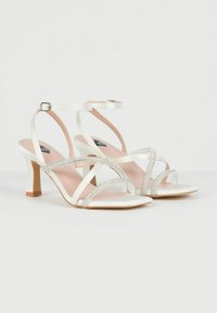 Sandales à talons hauts en satin blanc avec des brides croisées et des accents en strass. Bout carré, boucle à la cheville et un talon en bois léger.