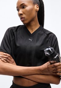 Γυναίκα με μαύρη αθλητική ενδυμασία Puma κρατάει μαύρο παγούρι, με τα χέρια σταυρωμένα, κοιτάζοντας προς τα πλάγια μπροστά σε απλό λευκό φόντο.