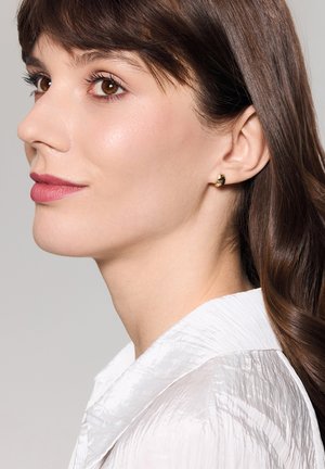 Złota kolczyk w kształcie obręczy z delikatnym żłobkowanym wzorem. Modelka ma na sobie białą koszulę o textured fakturze, z złożonym kołnierzykiem i gładkimi ciemnymi włosami.