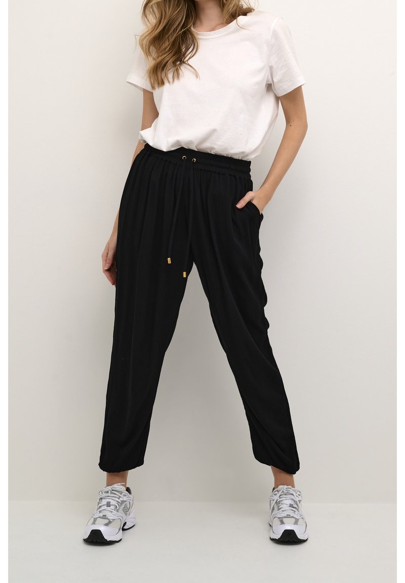 Femme portant un pantalon de jogging noir ample avec cordon de serrage et des baskets blanches, associé à un t-shirt blanc rentré, debout devant un fond uni.
