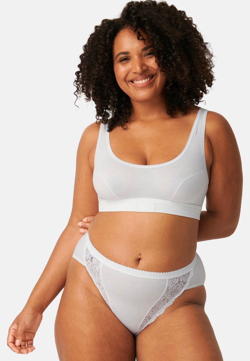 Femme souriante aux cheveux bouclés portant un soutien-gorge de sport blanc et un slip taille haute orné de dentelle, debout sur un fond uni.