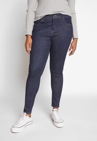 Persona con jeans skinny azul oscuro, un suéter de canalé gris claro y zapatillas blancas de caña baja, de pie frente a un fondo blanco sencillo.
