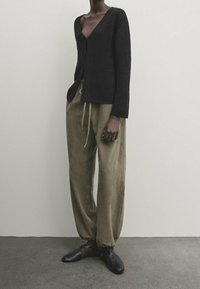 Schwarzer Strickcardigan mit V-Ausschnitt, getragen über lockeren, olivgrünen Hosen mit Kordelzugbund, kombiniert mit schwarzen Slippern.