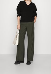 Czarny dzianinowy sweter z dekoltem w serek w połączeniu z oliwkowozielonymi szerokimi spodniami. Modelka trzyma kremową torebkę z teksturowanym wzorem i złotymi ozdobami.