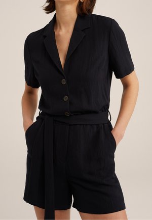 Kvinde iført sort kortærmet button-up romper med krave og stofbælte, hænderne i lommerne, neutral baggrund.