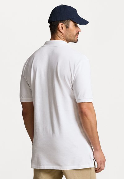 Polo shirt blanc en tissu texturé, à manches courtes et avec un ourlet arrondi. Associé à une casquette bleu marine, vu de dos.