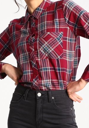 Kvinde iført en rød, blå og hvid ternet skjorte knappet op, stoppet ind i højttaljede sorte jeans, hænderne hvilende på hofterne.