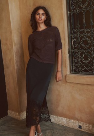 Fiatal nő sötét göndör hajjal, sötét kötött felsőt és hosszú csipkés szegélyű szoknyát visel, egy bézs, struktúrált falnak támaszkodik egy díszes ablakrács közelében.