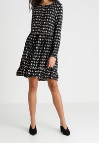 Černé šaty s dlouhým rukávem a přiléhavým korzetem, s rozšířenou sukní, které mají geometrický vzor v bílé, růžové a zelené barvě. Volánkový lem.