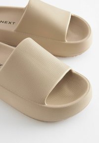 Beige slidesandaler lavet af tekstureret gummi, med åben tådesign og en forhøjet, mønstret sål for godt greb.