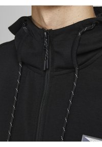 Primo piano di una felpa nera con zip, dotata di lacci riflettenti e di un logo parzialmente visibile sul petto.