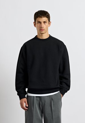 Ung mand med kort mørkt hår står iført en sort oversized sweatshirt og grå plisserede bukser mod en ensfarvet lys baggrund.