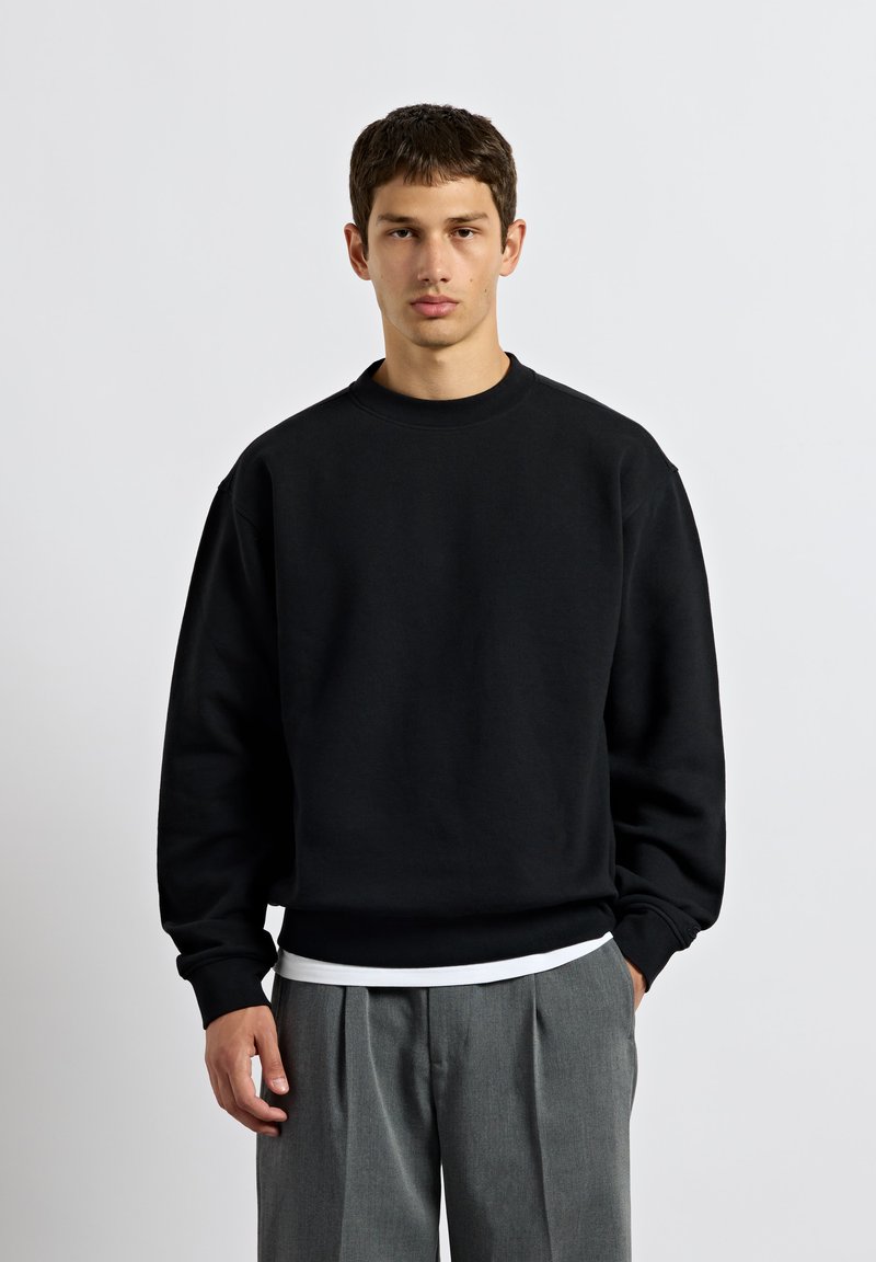 Junger Mann mit kurzen dunklen Haaren steht und trägt einen schwarzen übergroßen Pullover und graue plissierte Hosen vor einem schlichten hellen Hintergrund.