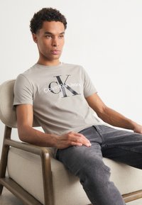 Pelēks kokvilnas T-krekls ar melnu "CK" logo un baltu "Calvin Klein Jeans" tekstu, pāris ar tumši pelēkiem džinsiem. Sēž uz bēša, polsterēta krēsla.