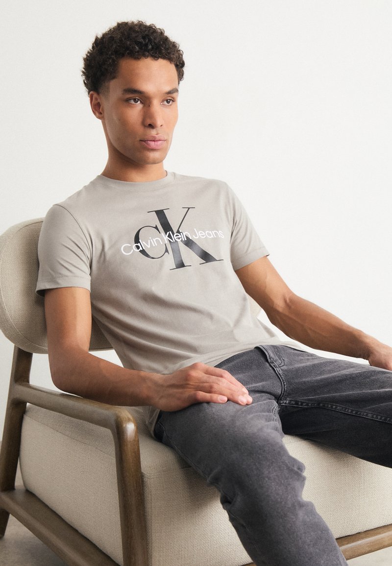 Pelēks kokvilnas T-krekls ar melnu "CK" logo un baltu "Calvin Klein Jeans" tekstu, pāris ar tumši pelēkiem džinsiem. Sēž uz bēša, polsterēta krēsla.