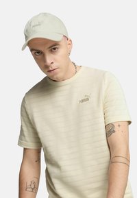 Puma lys beige stribet t-shirt med rund halsudskæring, korte ærmer og diskret logo. Suppleret med en matchende beige kasket.