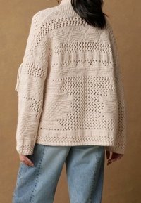Svetle béžový pletený svetr s otevřeným vzorem, se stojáčkem a texturovaným designem. Kombinován s volnými modrými džínami.