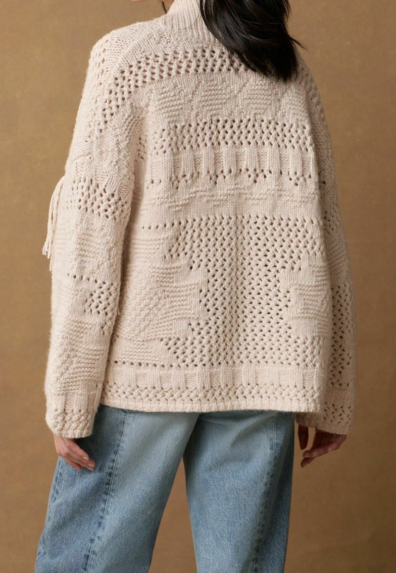 Svetle béžový pletený svetr s otevřeným vzorem, se stojáčkem a texturovaným designem. Kombinován s volnými modrými džínami.