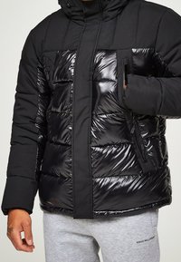 Svart uppblåsbar jacka med en glänsande yta, featuring en huva, med dragkedjefickor och ett kviltat mönster. Materialet är slätt och vadderat.