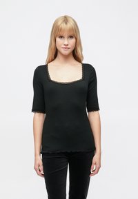 Musta toppi, jossa neliskanttinen pääntie, puolipitkät hihat ja aaltoileva pitsireuna. Valmistettu ribbineuloksesta, jossa on sileä pinta.