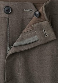 Tessuto marrone texture con una cerniera, chiusura con bottone e una tasca interna nascosta con cuciture grigie. Primo piano dei dettagli della vita.