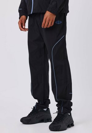 Persoon in zwarte trainingsbroek met blauwe bies en zwarte Nike sneakers, staand tegen een effen lichte achtergrond.