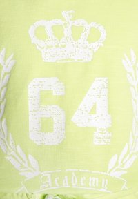 Limonkowa koszulka z białą koroną i liczbą "64" o zniszczonej teksturze, otoczona wzorem gałęzi oliwnej i słowem "Akademia."