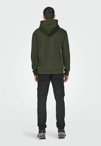 Felpa verde oliva con cappuccio regolabile, abbinata a pantaloni cargo neri. Le scarpe hanno accenti multicolori e suola robusta.