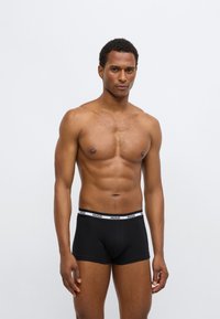 Μαύρα μπόξερ με λευκή ελαστική μέση, τυπωμένη με τη λέξη "HUGO." Το ύφασμα φαίνεται λείο και με εφαρμοστή σχεδίαση.