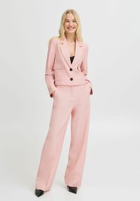 Světle růžový elegantní oblek s krčeným sakem, které má dvě přední kapsy a černé knoflíky, v kombinaci s kalhotami širokého střihu a špičatými černými lodičkami.